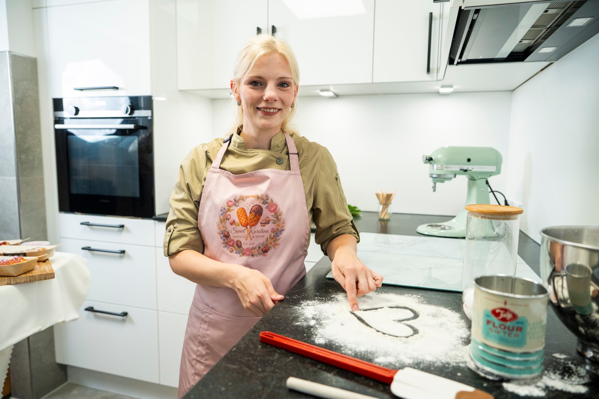 Karolina in ihrer Küche mit rosa Schürze und dem Sweet Karolina Logo, lächelnd am Arbeitstisch